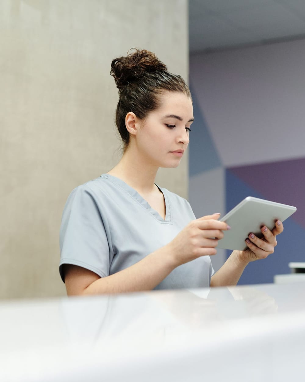 A receptionist in scrubs reading a tablet at a clinic front desk