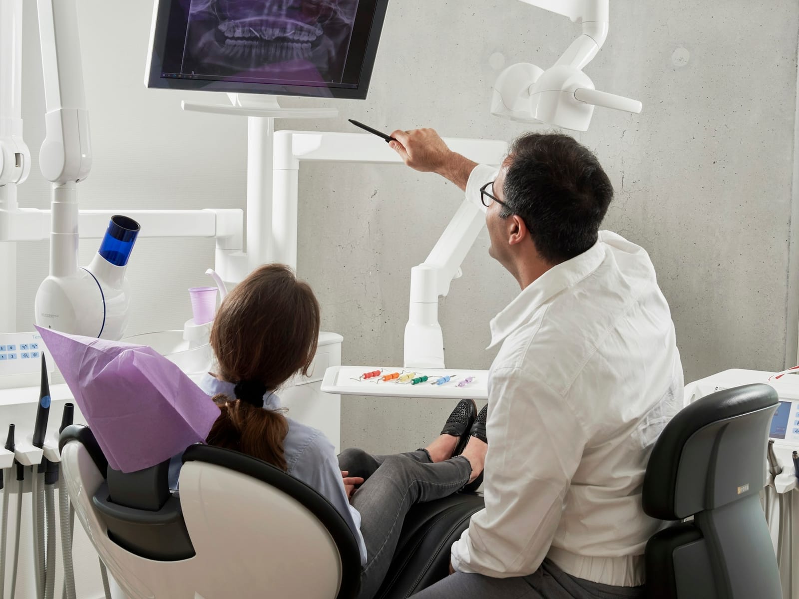 A dentist pointing at a dental X-ray on a screen while a patient looks on from the chair